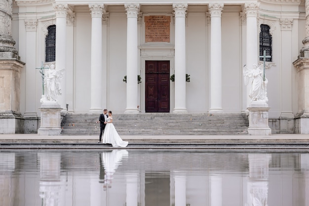 Tiny wedding couple is walking near the huge cathedral with white columns and reflection in the water