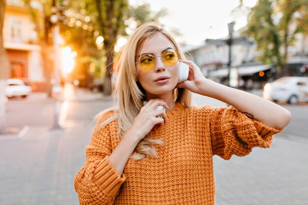 Усталая очаровательная дама в оранжевом свитере стоит на городской площади и слушает любимую песню в наушниках