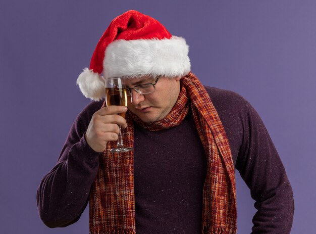 보라색 배경에 고립 된 닫힌 눈을 가진 샴페인 잔으로 이마를 만지고 목 주위에 스카프와 안경과 산타 모자를 쓰고 피곤 성인 남자