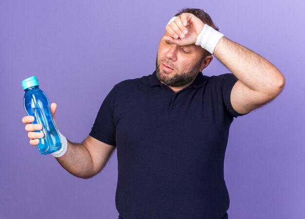 усталый взрослый славянский спортивный мужчина с повязкой на голову и браслетами, положив руку на лоб и держа бутылку с водой, изолированную на фиолетовой стене с копией пространства
