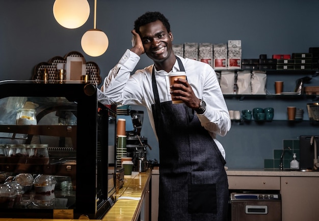 Усталый африканский бариста держит чашку с кофе, опираясь на прилавок в кафе и глядя в камеру со счастливым взглядом.