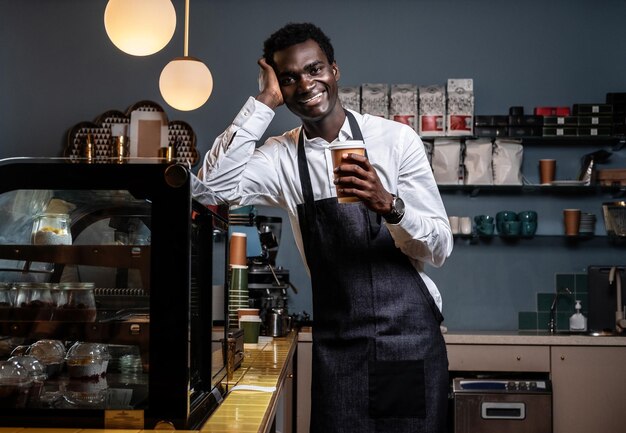 Tired African barista holding a cup with coffee while leaning on a counter in a coffee shop and looking at a camera with a happy look .