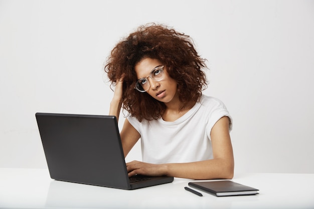 Lavoro di pensiero della donna di affari africana stanca al computer portatile sopra la parete bianca.