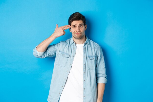 Tired and annoyed adult man making finger gun gesture near head, shooting himself from boredom, standing against blue background