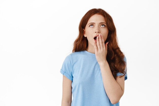 Tired and annoyed redhead girl, yawning and roll eyes uninterested, standing sleepy and unamused, hear boring story, standing against white background.