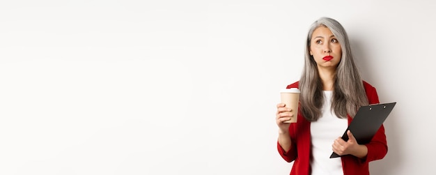 Free photo tired asian female office worker holding clipboard and paper cup drinking coffee and exhaling with e