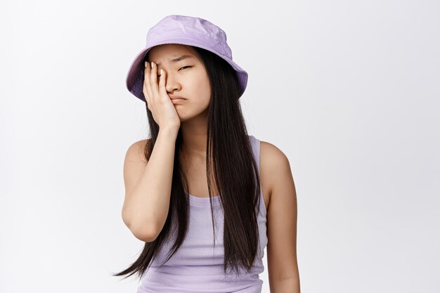 疲れたアジアの女の子のfacepalmは、白い背景に立っている何かに悩まされてため息をつき、疲れ果てているように見えます