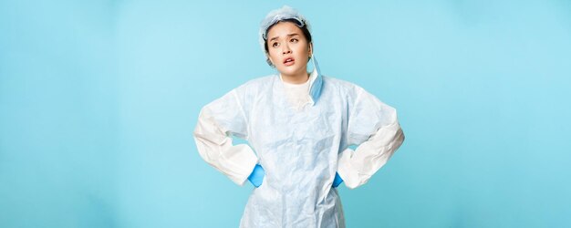 Tired asian nurse or doctor take off medical mask feeling fatigue wearing personal protective equipment sighing with hands on waist blue background