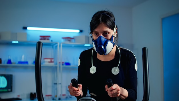 Free photo tired athlete woman training on cross trainer increasing rhythm of exercises wearing mask and medical electrodes monitoring muscle endurance and cardiac rate in science sport laboratory