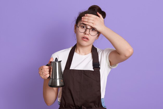 Tired attractive teenage girl wearing white t shirt and brown apron, holding tea or coffee pot in hands