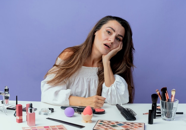 Tired beautiful girl sits at table with makeup tools puts hand on chin looking isolated on purple wall
