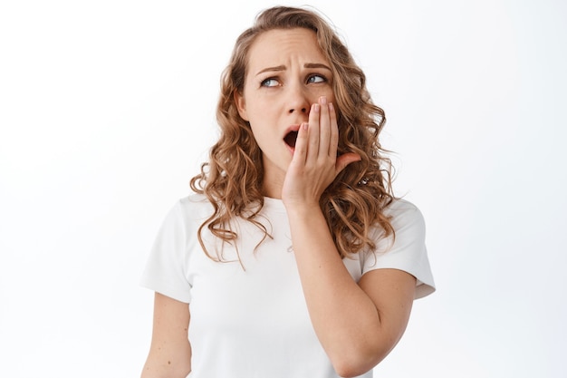 Ragazza bionda stanca che si sente assonnata o annoiata, guardando da parte il testo promozionale con fatica, fissando qualcosa di noioso, in piedi sul muro bianco