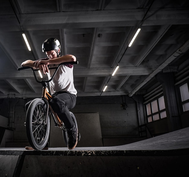 Tired BMX rider in protective helmet sitting on his bicycle in a skatepark indoors