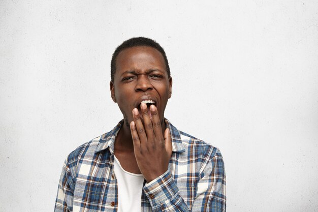 Усталый или скучающий афро-американский юноша прикрывает рот, зевая, чувствуя себя измотанным после тяжелого рабочего дня. Чернокожий студент, имеющий сонный скучный взгляд во время урока истории в колледже