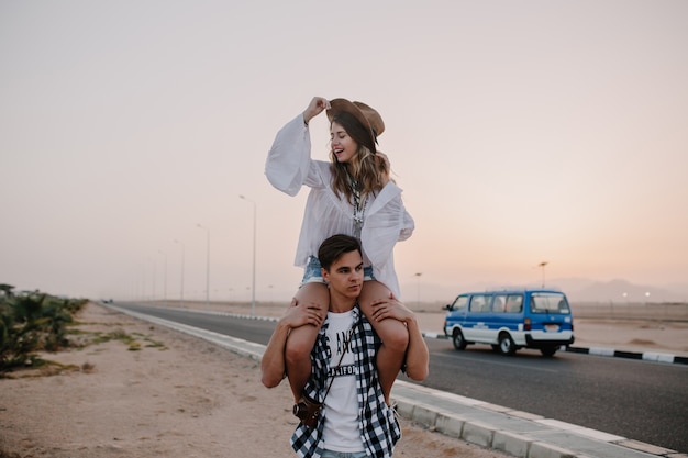 Tired brunette guy in plaid shirt carries his girlfriend on his shoulders looking at sunset. Cheerful young woman in trendy hat having fun with boyfriend on outdoor evening date