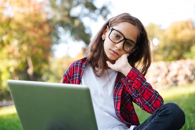 Tired brunette schoolgirl in eyeglasses sitting on grass