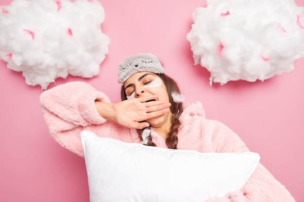 Tired brunette woman with closed eyes covers mouth with hand wears sleepmask and pajama