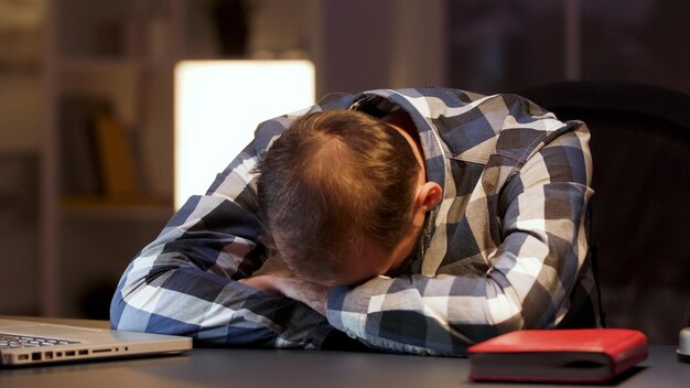 ホームオフィスの机の上で頭を抱えて寝ている疲れたビジネスマン。働き過ぎの起業家。