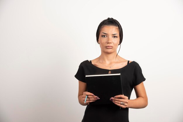 Tired businesswoman posing with notebook on white wall.