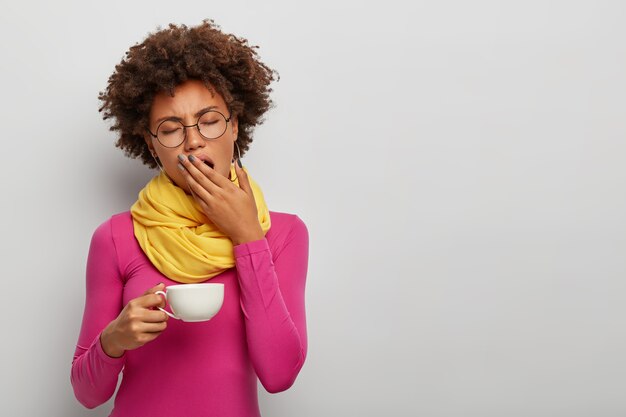 疲れた巻き毛の女性のあくび、眠そうな表情、早朝にコーヒーを飲む、熱い飲み物の白いマグカップを保持