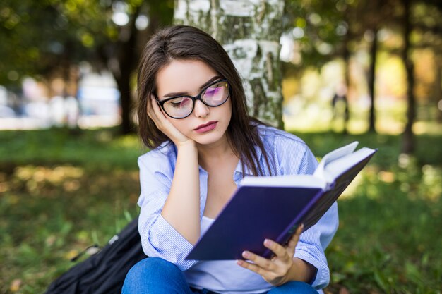 Tired dark-haired serious girl in jeans jacket and glasses reads book against summer green park.