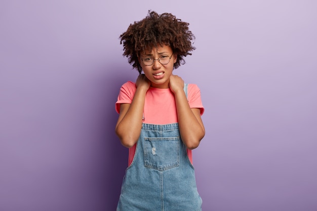 Free photo tired displeased cheerless woman has afro hair keeps both hands on neck, smirks face from pain, being tired of working at computer