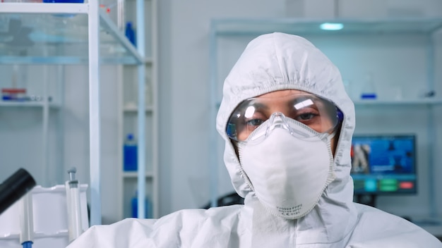 Tired doctor wearing coverall looking exhausted at camera in modern equipped lab. Scientist examining virus evolution using high tech and chemistry tools for scientific research, vaccine development.