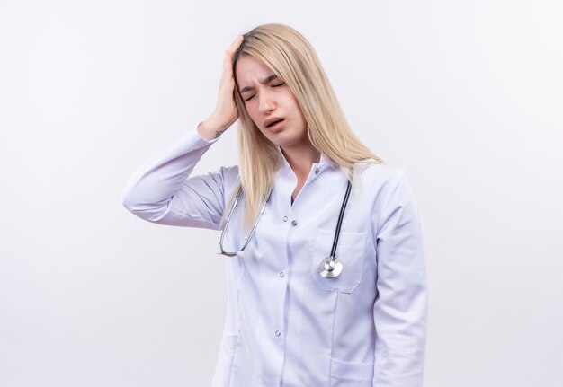 Tired doctor young blonde girl wearing stethoscope and medical gown put her hand on head on isolated white wall