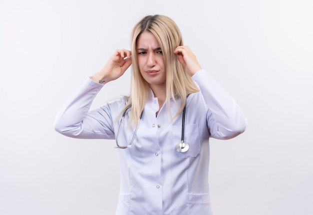 Tired doctor young blonde girl wearing stethoscope and medical gown put her hands on head on isolated white wall
