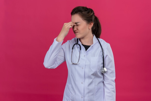 Усталая девушка-врач в медицинском халате и стетоскопе положила руку на лоб на изолированном красном фоне