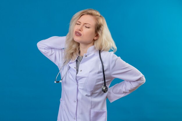 Tired doctor young girl wearing stethoscope in medical gown put her hand on neck on blue background