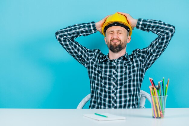 Tired engineer man is putting hands behind head on blue background