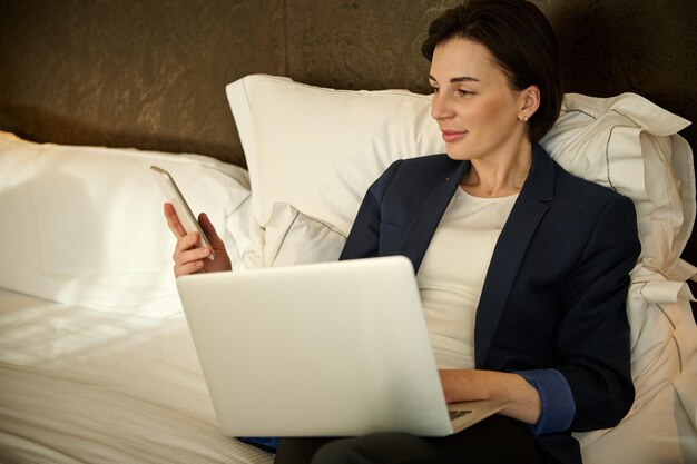 피곤한 유럽 젊은 비즈니스 여성은 침대에 누워서 휴대전화와 노트북 컴퓨터를 사용하고 출장에서 비즈니스 파트너와 비즈니스 미팅을 한 후 호텔 방에서 휴식을 취합니다.
