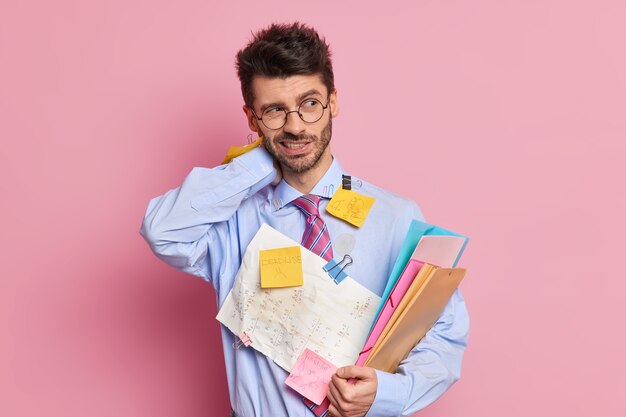 Tired exhausted employee has neckpain holds folders and wears shirt with attached sticky notes written information poses 