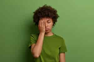 Free photo tired exhausted woman makes face palm, has problems with health, sleepy displeased expression, sighs from tiredness, wears casual green t shirt, poses indoor. feeling fatigued and overworked