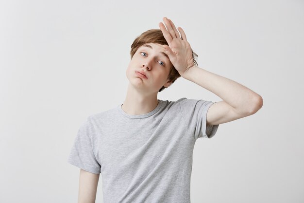 Tired fair-haired young man feels headahce after hard work, touches head with hand, stressed. Regretful caucasian guy has forgetful expression, tries to remember all necessary details