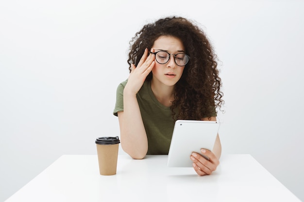 Tired fed up girlfriend with curly hair, sitting at table in cafe, drinking tea or coffee and reading article in digital tablet, holding hand on temple