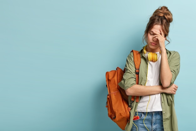 Tired female student has sleepy expression, tries to refresh herself with music in headphones, carries rucksack, wears casual clothes