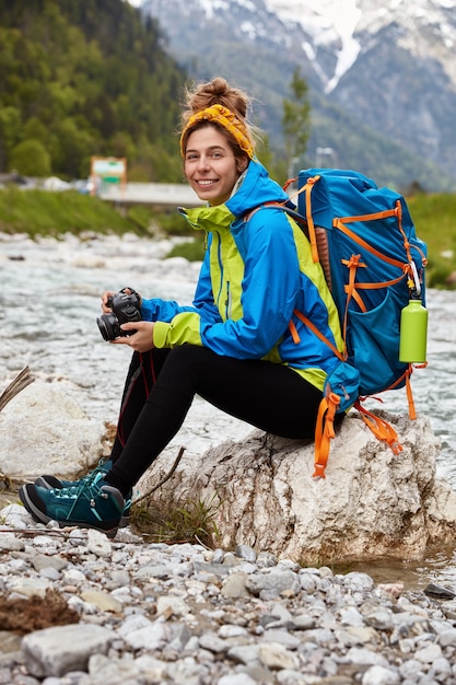 Tired female tourist sits on stones near little stream in mountains, holds professional camera, views photos