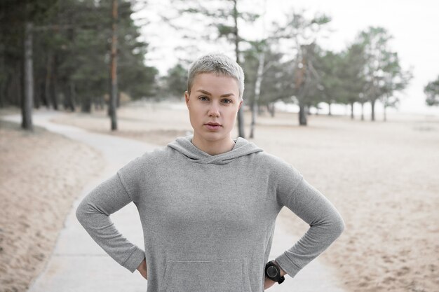 Tired fit slim girl in hoodie posing outdoors, keeping hands on her waist and looking at camera, having rest during cardio running workout. People, lifestyle, activity, health and fitness concept