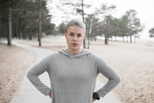 Tired fit slim girl in hoodie posing outdoors, keeping hands on her waist and looking at camera, having rest during cardio running workout. People, lifestyle, activity, health and fitness concept