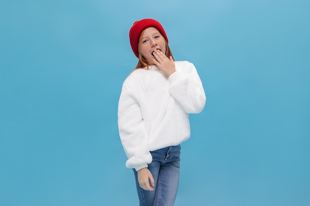 Tired ginger girl with freckles in oversize shirt and jeans yawns on blue background Young cute lady in red hat posing on isolated backdrop