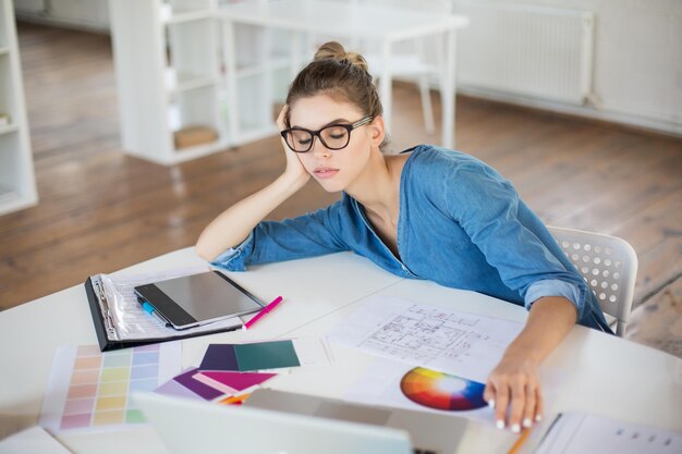 Tired girl in denim shirt and eyeglasses leaning on hand sleeping while spending time in modern office