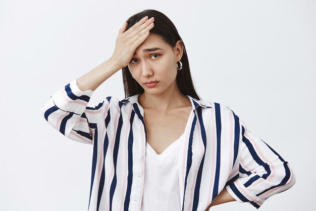 Tired and gloomy cute woman in striped blouse, holding palm on forehead, feeling under pressure, being exhausted and gazing upset over gray wall