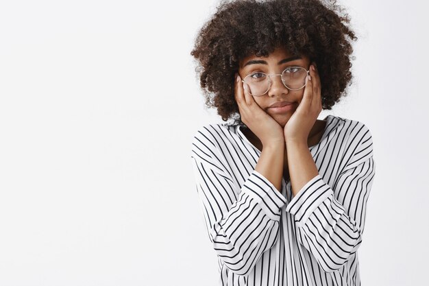 tired and gloomy indifferent dark-skinng young girl with afro hairstyle squeezing face with palms on cheeks staring careless and exhausted dreaming about day off