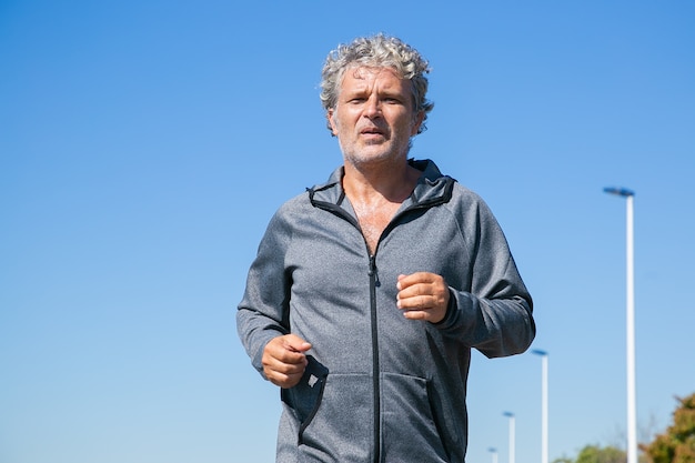 Tired grey haired man in sports jacket jogging outside. Senior jogger training in morning. Front view, blue clear sky, copy space. Activity and age concept