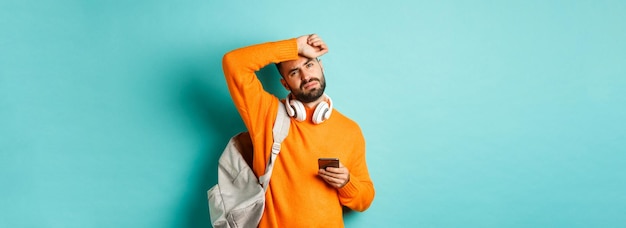 Free photo tired guy with headphones and backpack wiping sweat off forehead with exhausted face using mobile ph