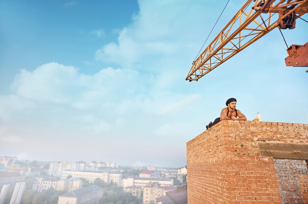 Tired and handsome builder in hat lying on brick wall on high and resting. Looking away. Blue sky with clouds at summer season on background. Milk and bread near.