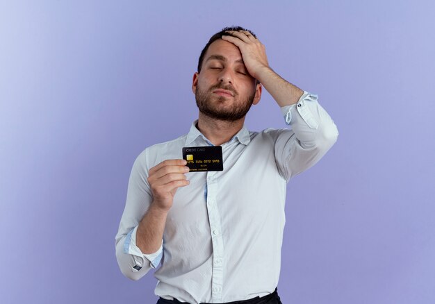 疲れたハンサムな男が紫色の壁に分離されたクレジットカードを保持している額に手を置きます。