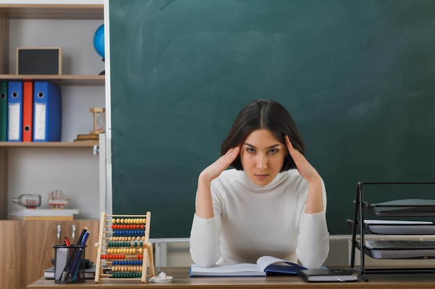 Free photo tired looking at camera young female teacher sitting at desk with school tools on in classroom